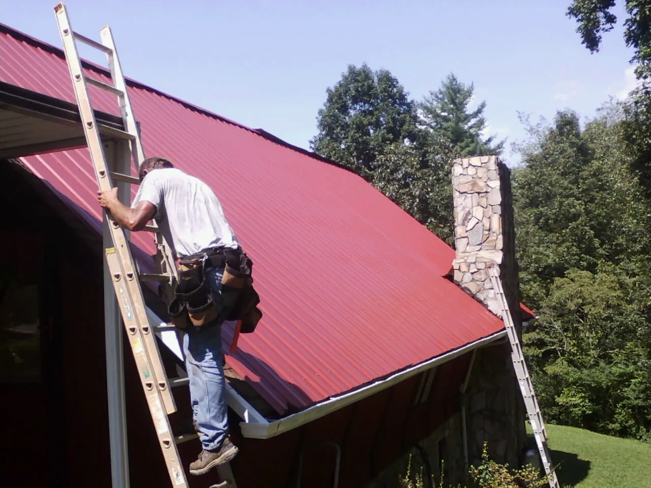 Roofer on a ladder beside a red metal roof and stone chimney — Soffit & Fascia Repair by Roofing Brothers in Wingina